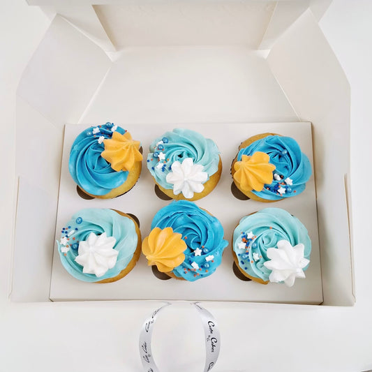 A box of beautifully decorated cupcakes to match the famous with light blue and dark blue rosettes, orange and white meringues, and sprinkles. Edible glitter is dusted on top.