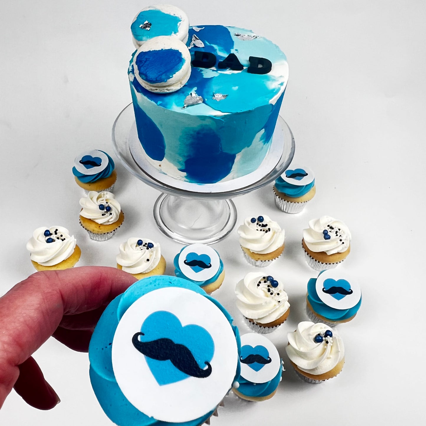 A blue and white watercolor-themed 'Dad' cake on a glass stand, surrounded by matching blue and white cupcakes with heart and mustache decorations. A hand is holding a cupcake in the foreground, set against a clean white background.