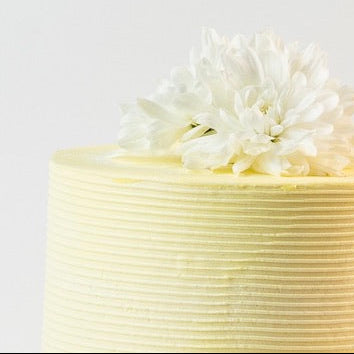 Close up image of yellow cake, decorated with ribbed buttercream feature on the sides and white fresh flowers on the top. Made with love by Brisbane’s favourite, 5 star cake shop, Cute Cakes & Co