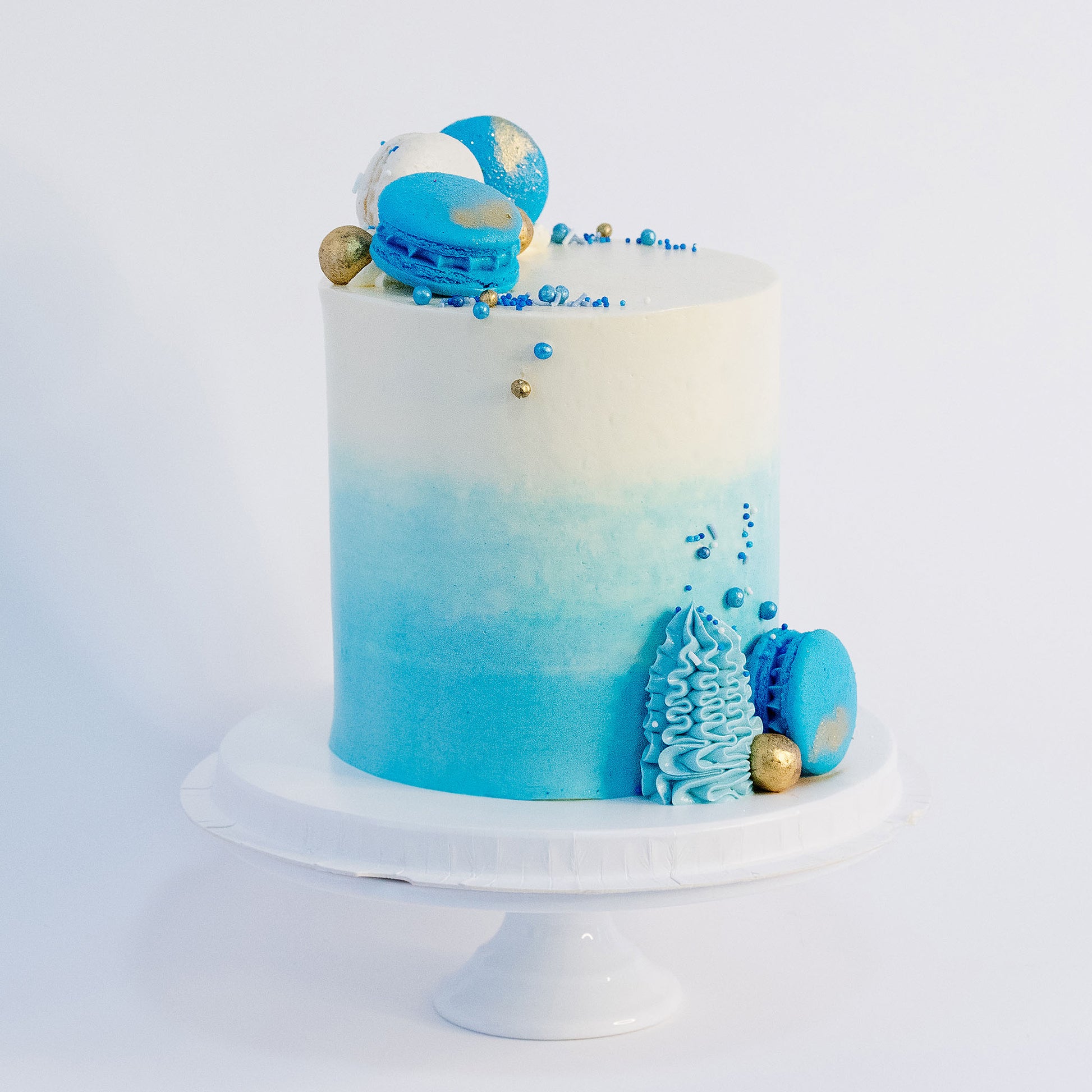 A beautifully decorated cake with ombre blue frosting- buttercream, topped with white and blue macarons, gold maltesers, and a medley of blue, white, and gold sprinkles.Made be Cute Cakes and Co. Brisbane Cakes shop near you