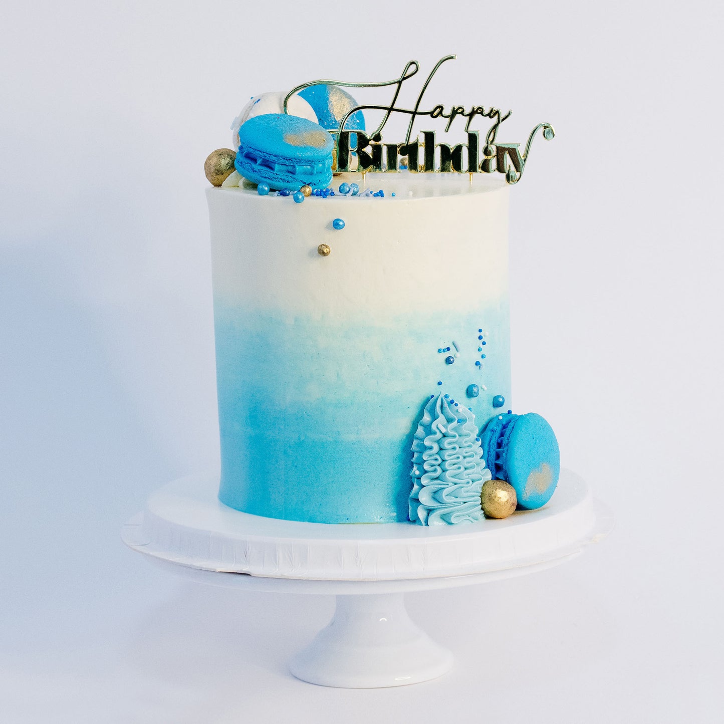 A beautifully decorated brithday cake cake with ombre blue frosting- buttercream, topped with white and blue macarons, gold maltesers, and a medley of blue, white, and gold sprinkles.Made be Cute Cakes and Co. Brisbane Cakes shop near you