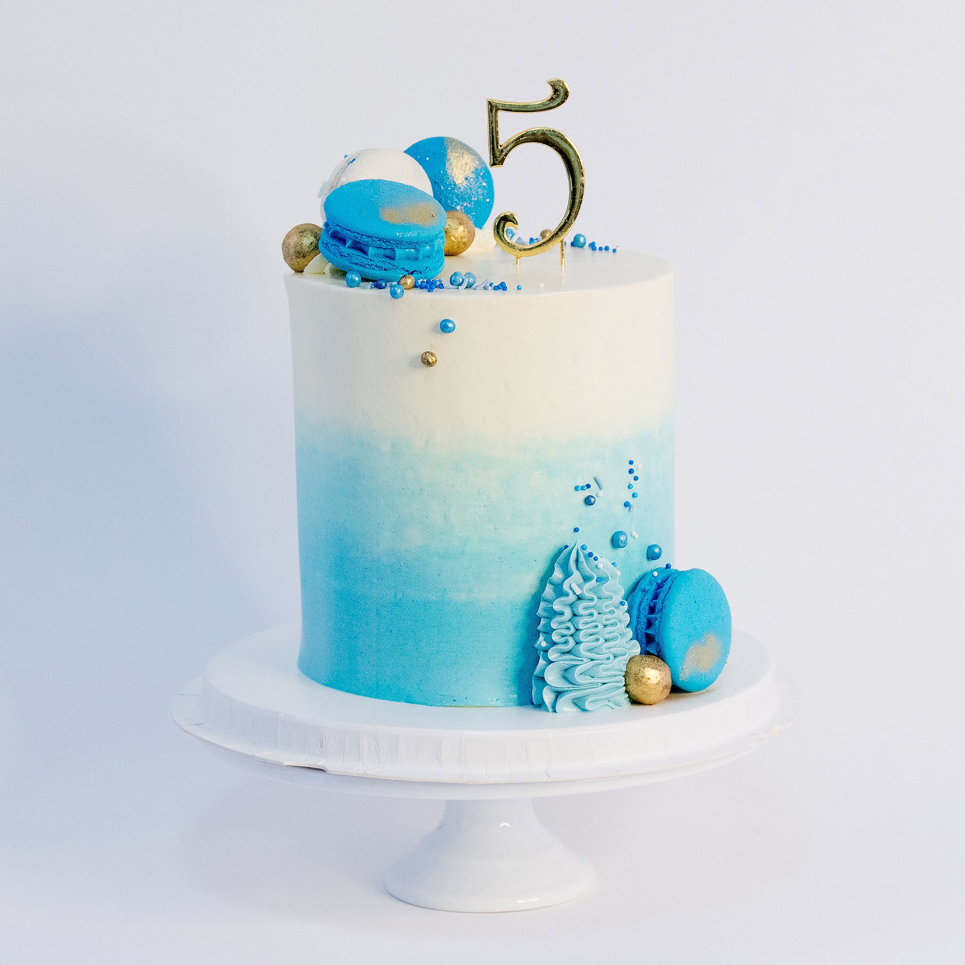 A beautifully decorated birthday cake with ombre blue frosting- buttercream, topped with white and blue macarons, gold maltesers, and a medley of blue, white, and gold sprinkles.Made be Cute Cakes and Co. Brisbane Cakes shop near you