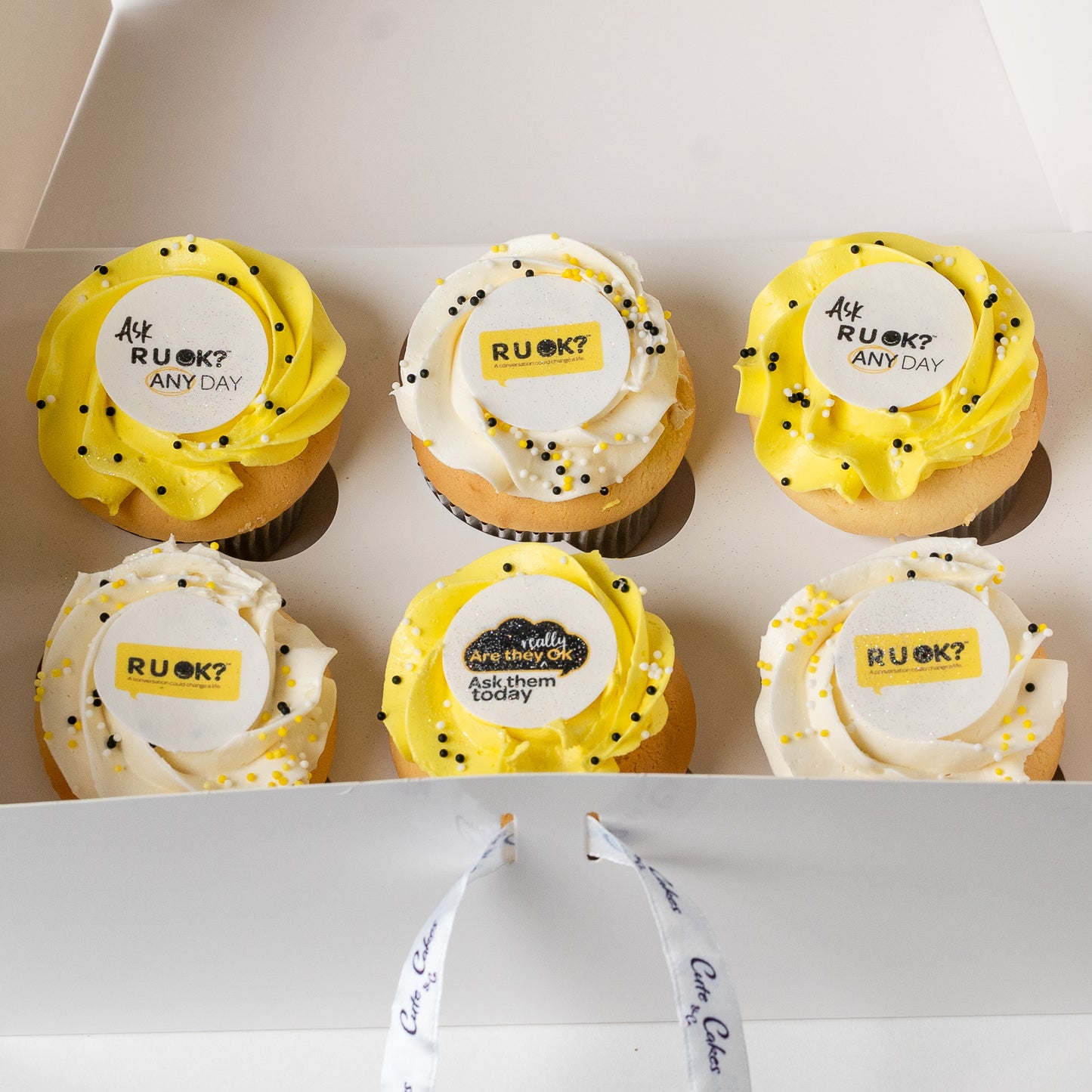Six cupcakes arranged in a white gift box, each topped with yellow or white frosting, black and gold sprinkles, and a round custom topper that reads 'Ask RUOK? ANY DAY'.