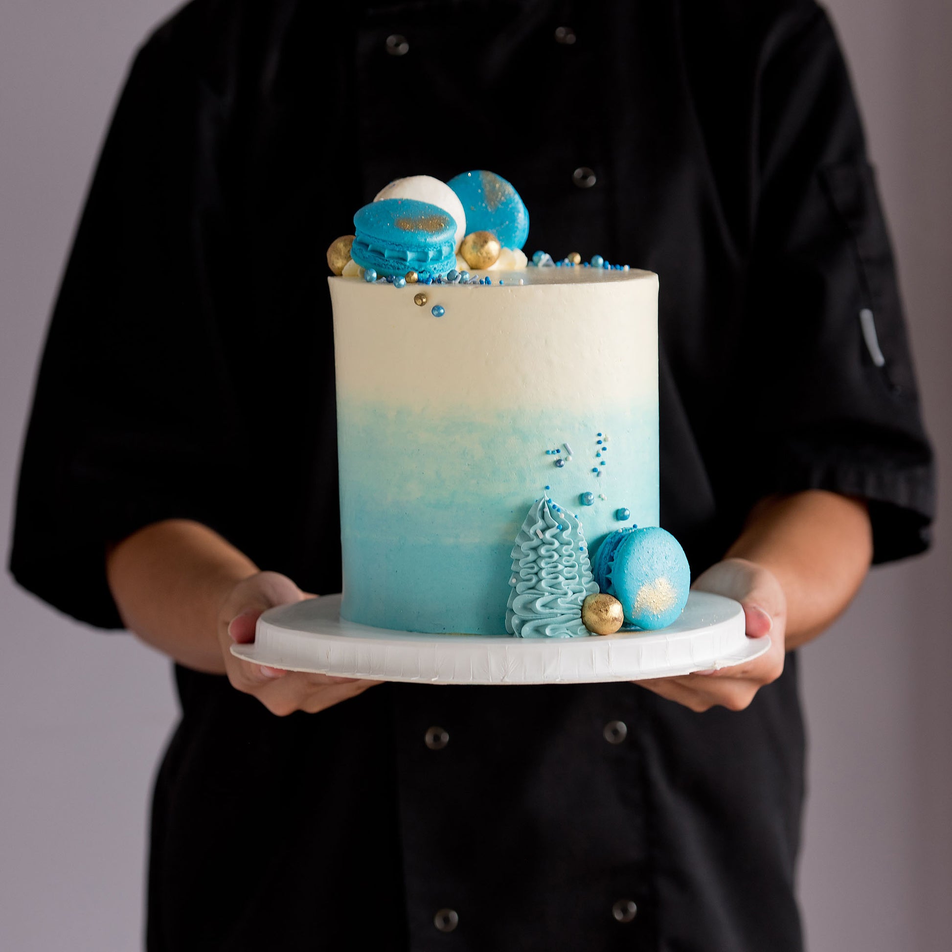 A beautifully decorated cake with ombre blue frosting- buttercream, topped with white and blue macarons, gold maltesers, and a medley of blue, white, and gold sprinkles.Made be Cute Cakes and Co. Brisbane Cakes shop near you
