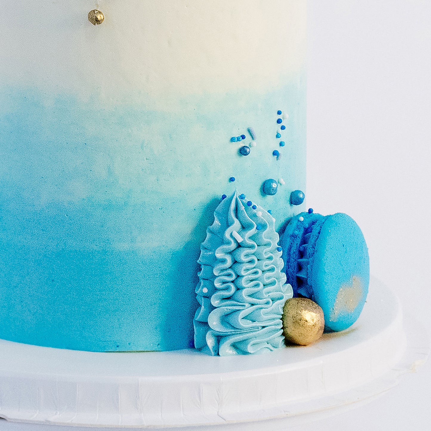 A beautifully decorated cake with ombre blue frosting- buttercream, topped with white and blue macarons, gold maltesers, and a medley of blue, white, and gold sprinkles.Made be Cute Cakes and Co. Brisbane Cakes shop near you