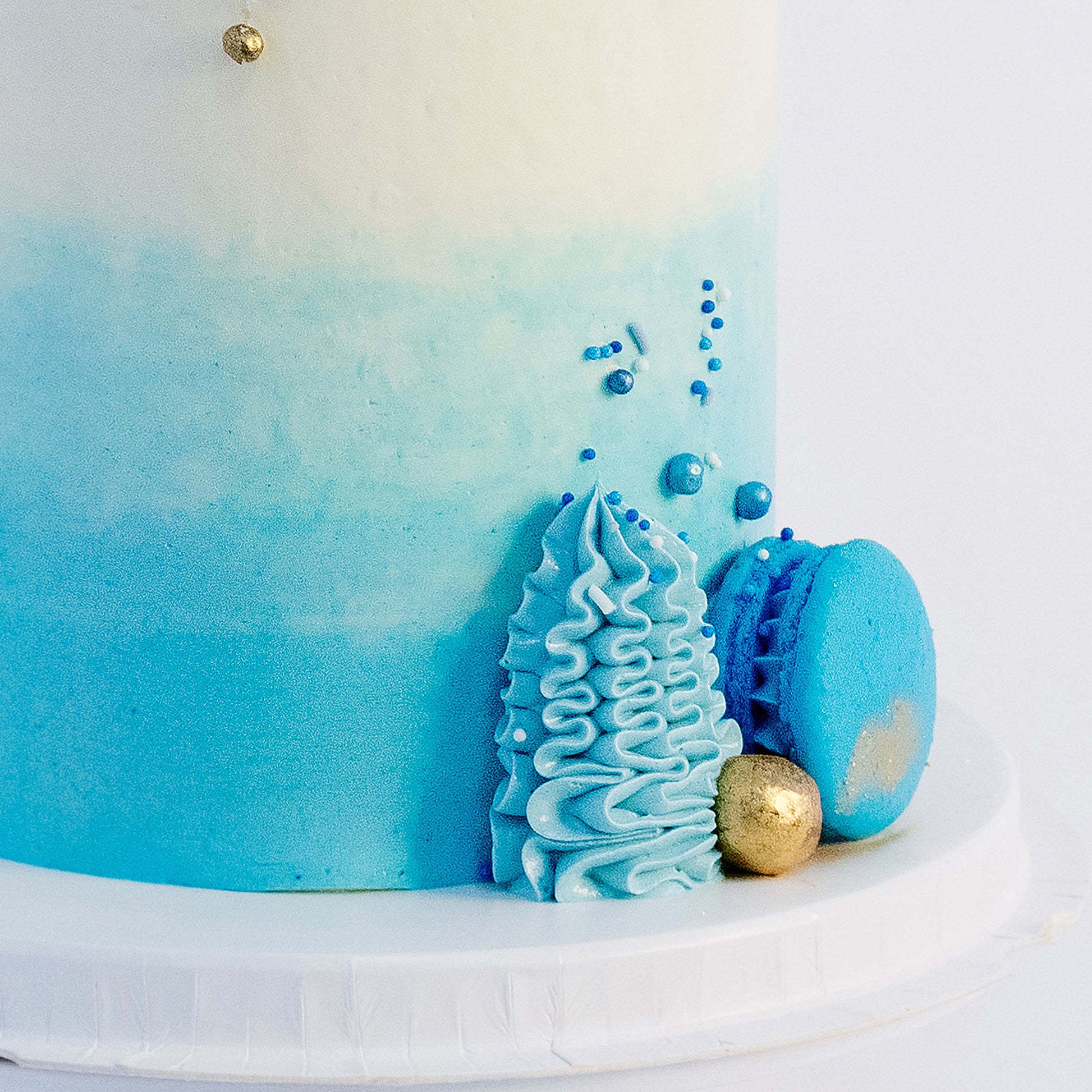 A beautifully decorated cake with ombre blue frosting- buttercream, topped with white and blue macarons, gold maltesers, and a medley of blue, white, and gold sprinkles.Made be Cute Cakes and Co. Brisbane Cakes shop near you