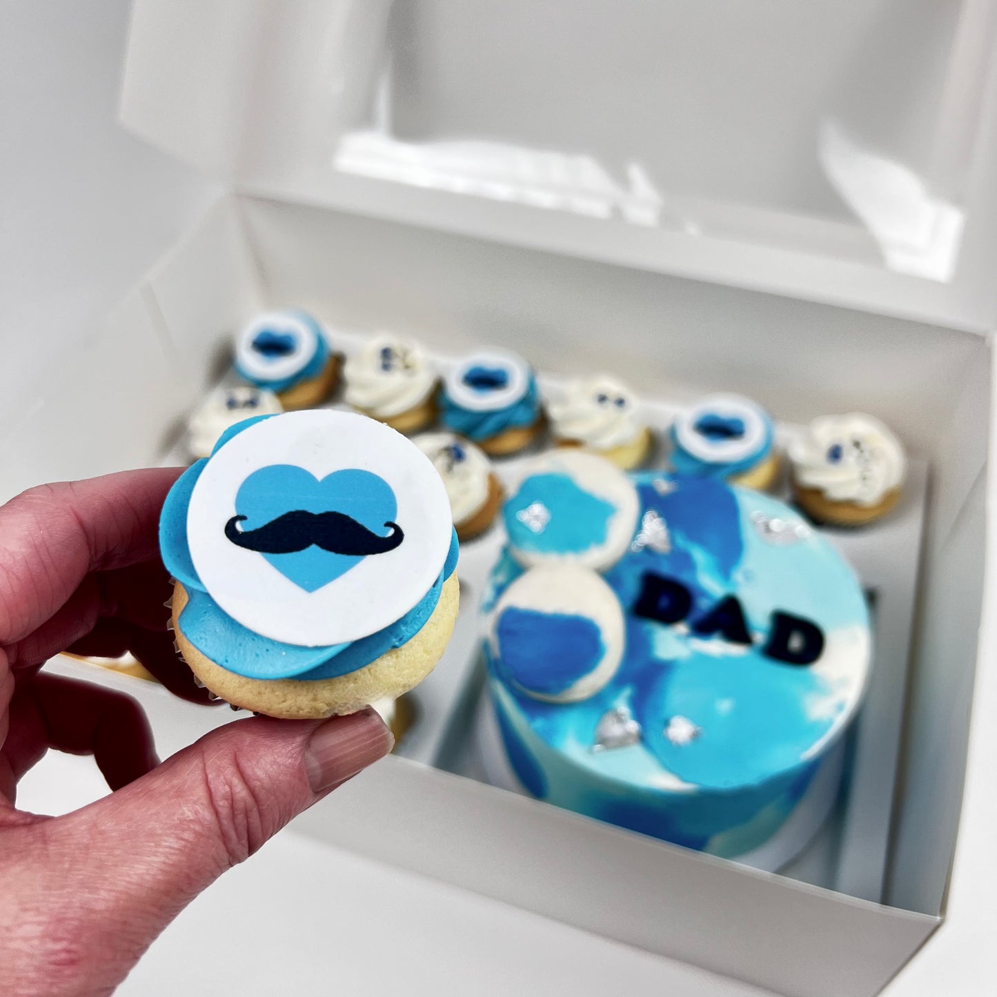 A hand holds a decorated cupcake in front of a white gift box containing a blue and white 'DAD' cake and matching cupcakes.