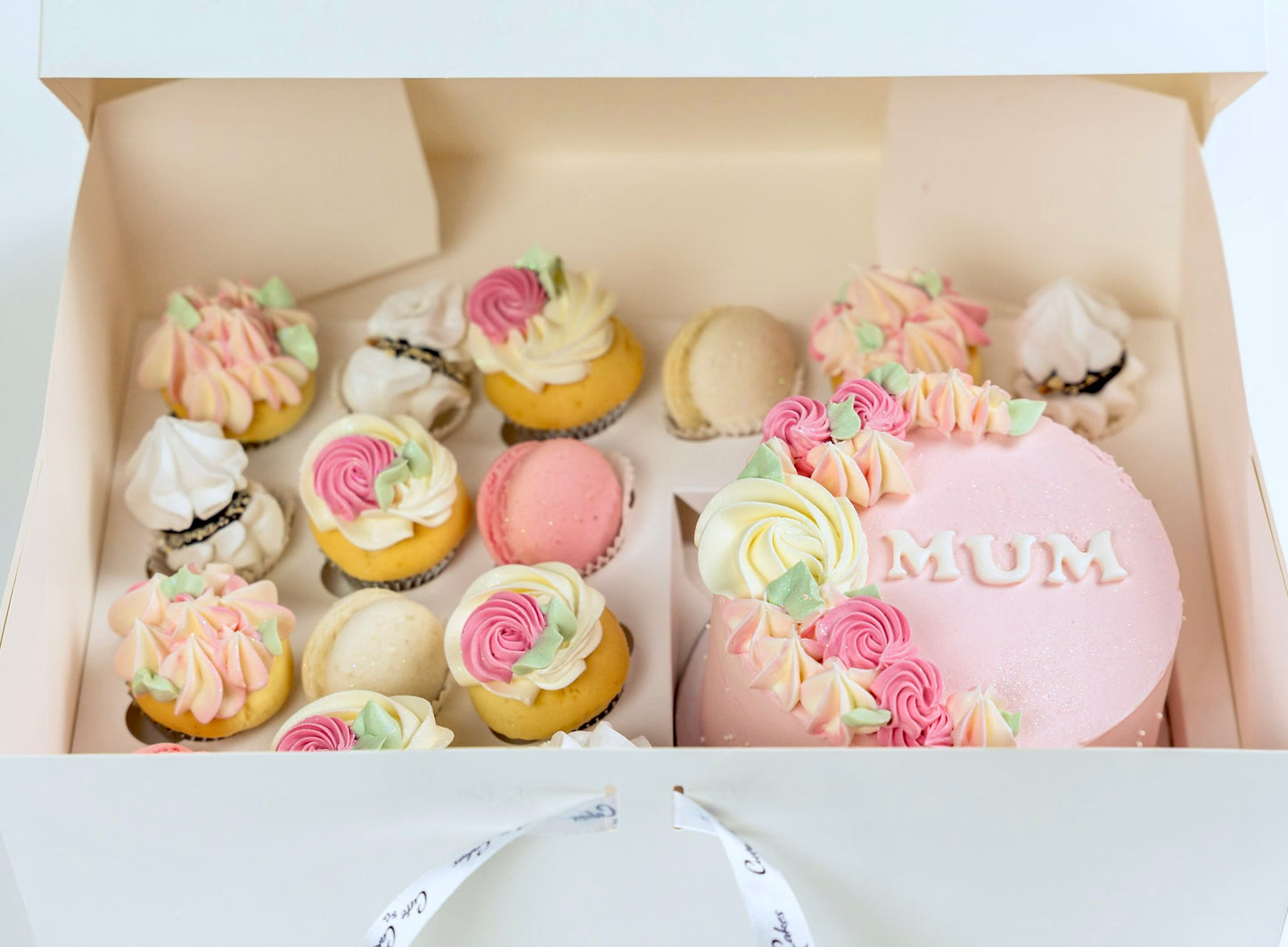 Decorative cake with 'MUM' on top, surrounded by smaller pastel-colored cakes in a box.