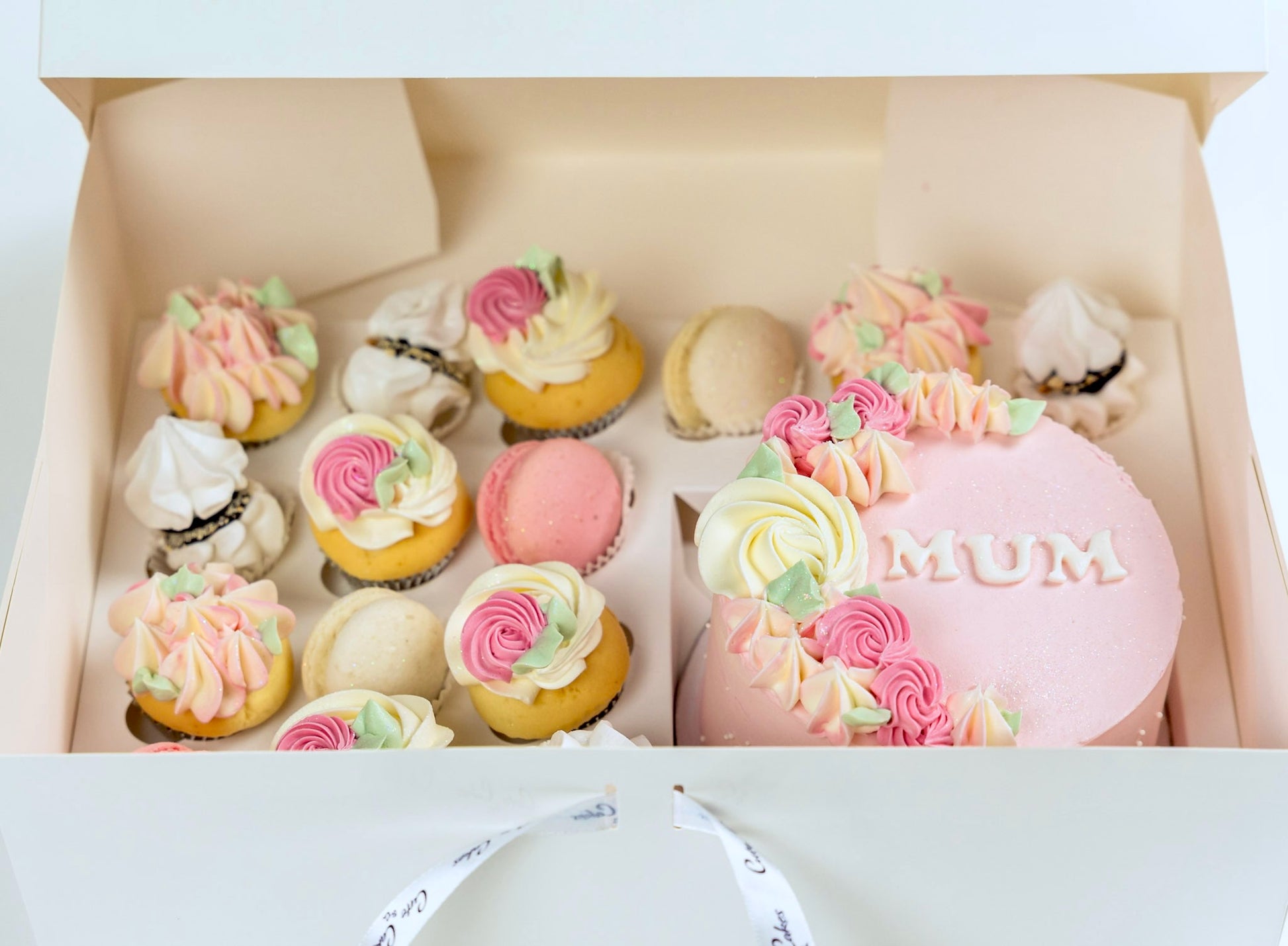 Decorative cake with 'MUM' on top, surrounded by smaller pastel-colored cakes in a box.