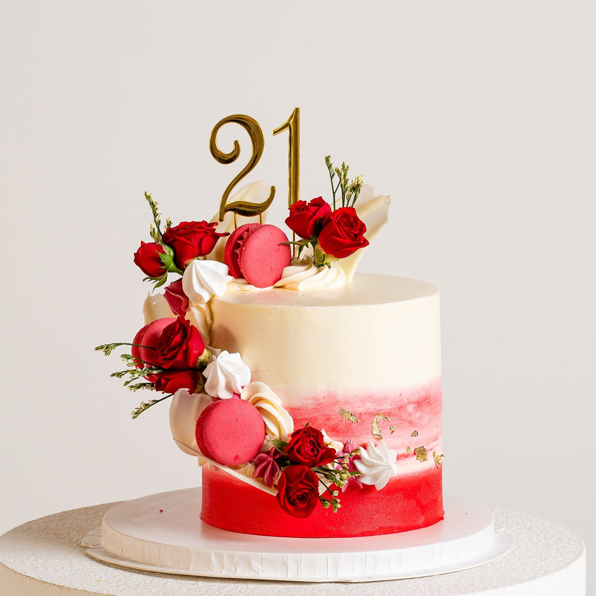 A decorated white buttercream birthday cake featuring ombre red bottom half with  red roses, red macarons, and a gold '21' topper.