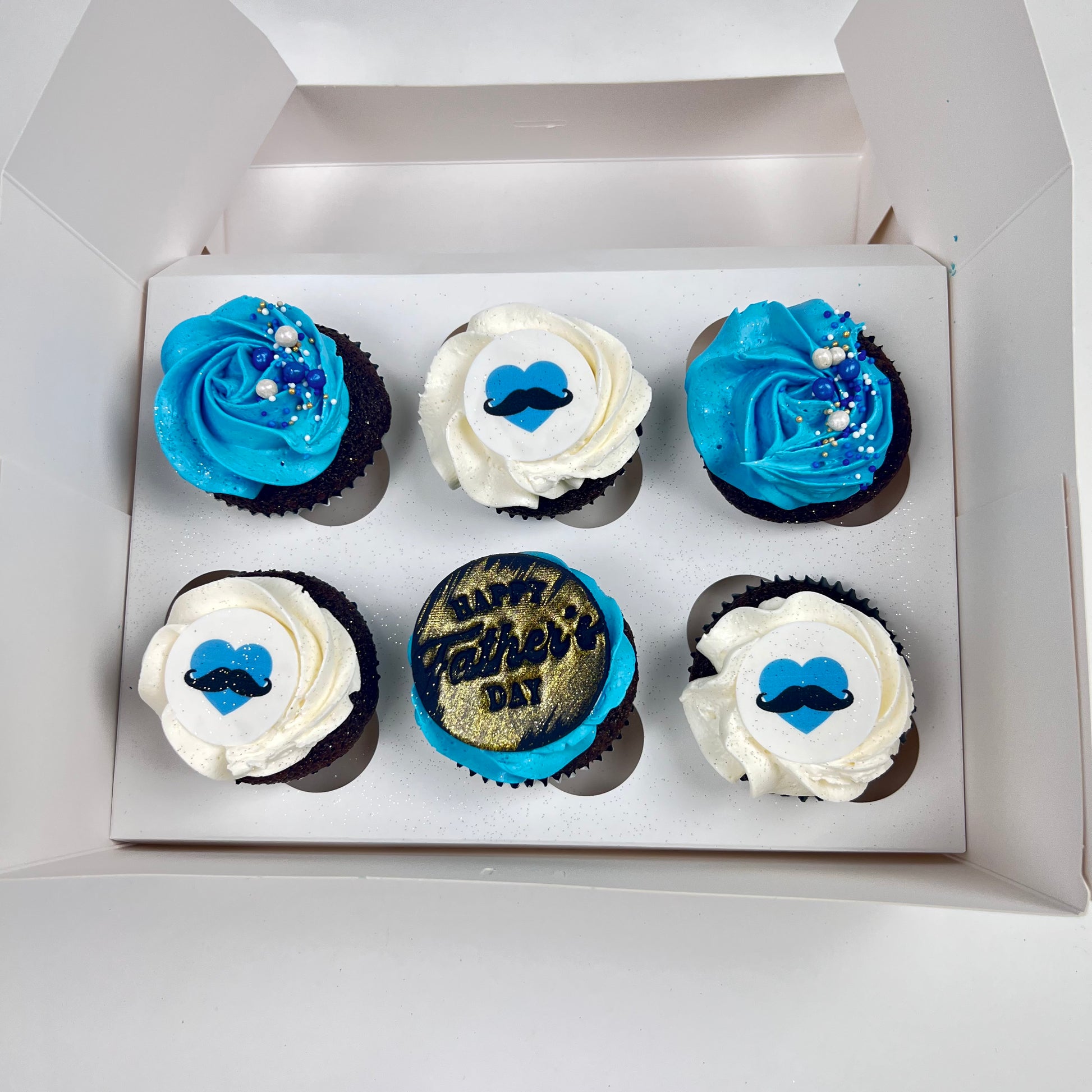 6 pack of blue and white decorated cupcakes. Half have a disk with a blue heart with a black moustache. one has Black plaque with Happy Father's Dad plaque . 