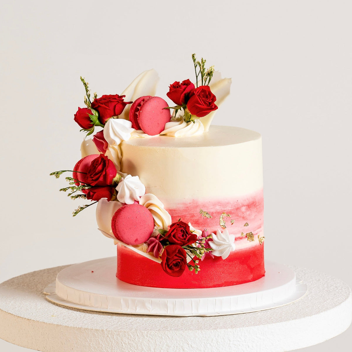 Front view of a red buttercream cake with cascade of macarons, roses, white chocolate sails and gold leaf. 