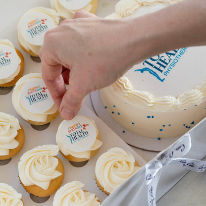 A hand is placing a custom round cupcake topper onto a mini cupcake. The edible topper with  company logo. white background with blue and red text 