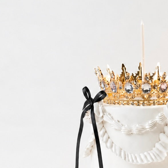 A lit candle on a white Vintage style birthday cake. Black ribbon bow with a gold crown on the top 