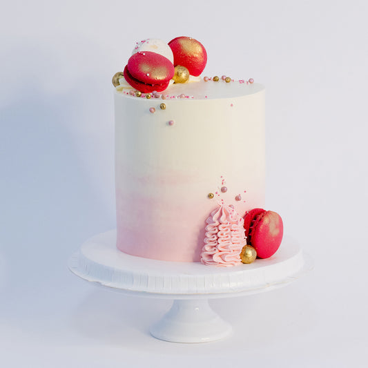 A beautifully decorated cake with a gradient of pink to white on the outside, topped with white and pink macarons, gold maltesers, and pink, white, and gold sprinkles. Made by Cute Cakes & Co . Brisbane cake shop that delivers the same day. 