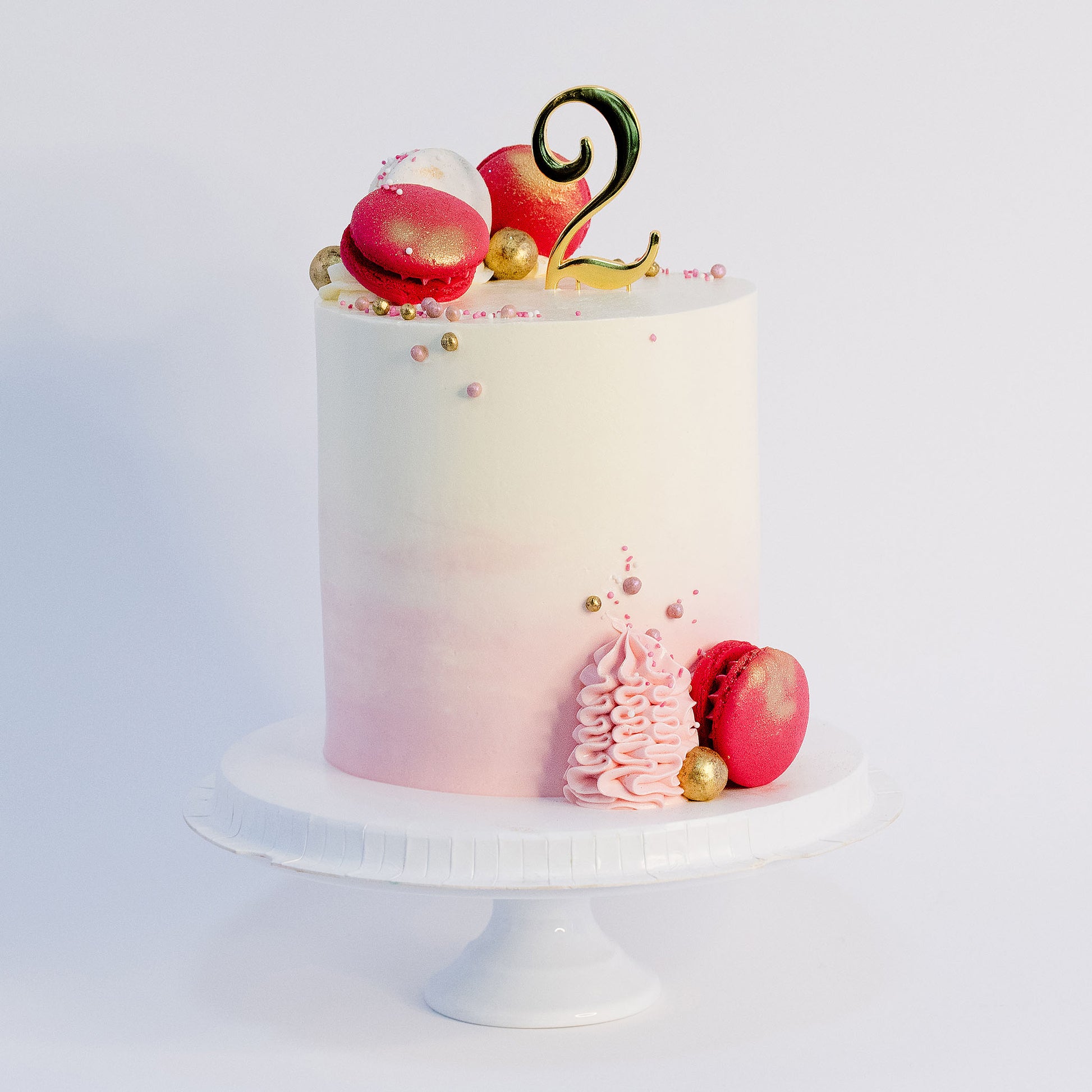 A beautifully decorated 2nd birthday cake with  pink and white macarons, gold maltesers, and pink, white, and gold sprinkles. Made by Cute Cakes & Co . Brisbane 5 star rated cake shop that delivers the same day. 