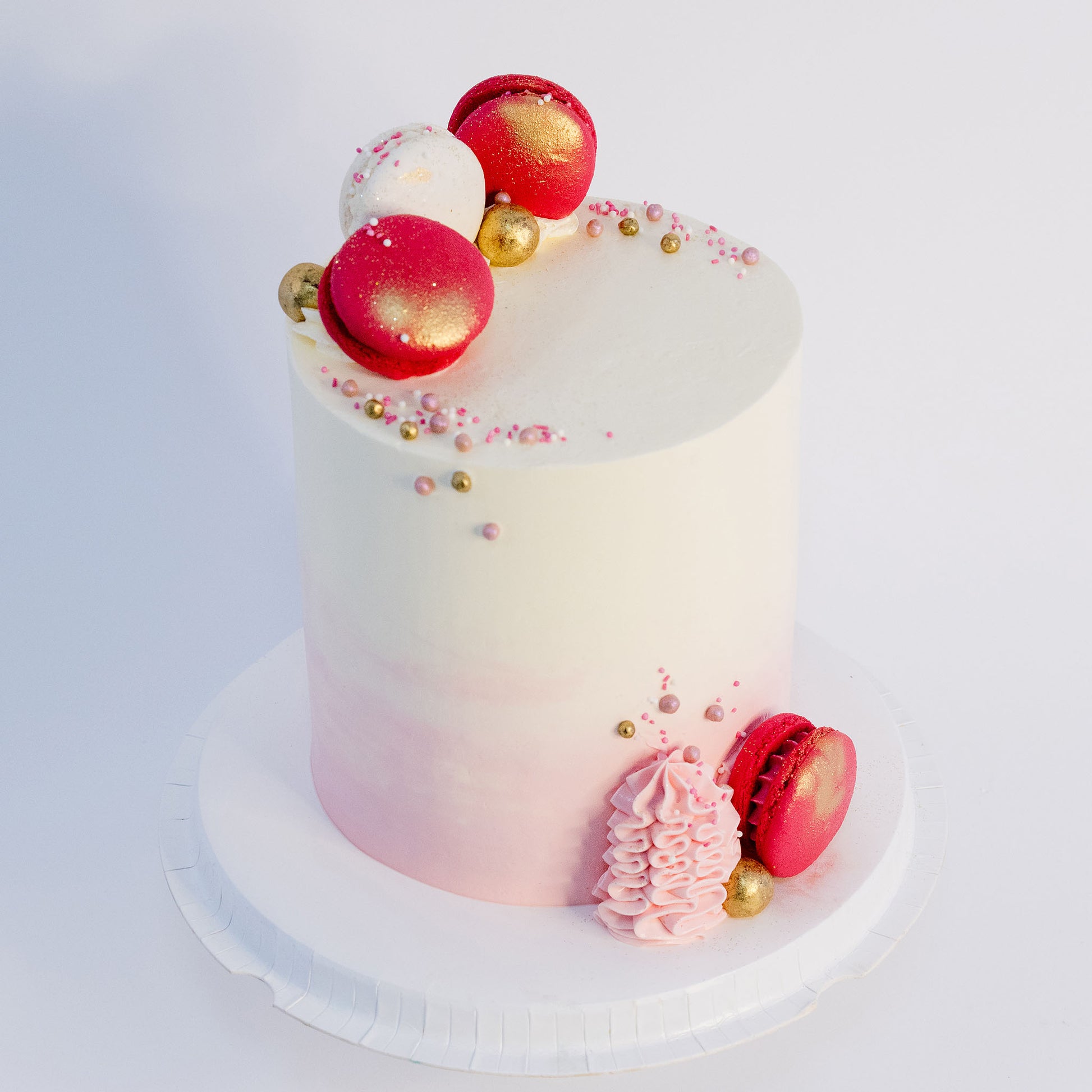 A beautifully decorated birthday cake with a gradient of pink to white on the outside, topped with white and pink macarons, gold maltesers, and pink, white, and gold sprinkles. Made by Cute Cakes & Co . Brisbane cake shop that delivers the same day. 