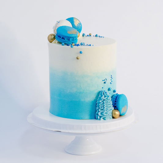 A beautifully decorated cake with ombre blue frosting- buttercream, topped with white and blue macarons, gold maltesers, and a medley of blue, white, and gold sprinkles.Made be Cute Cakes and Co. Brisbane Cakes shop near you 