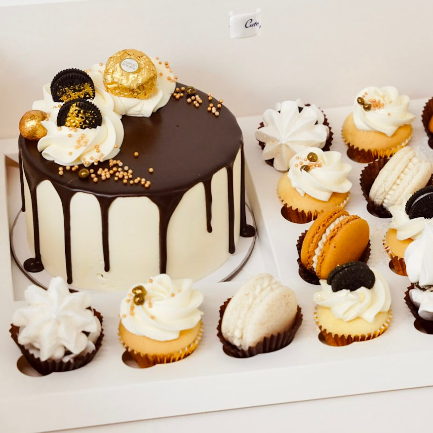 Chocolate cake with drizzle and Ferrero Rocher on top, surrounded by cupcakes and cookies on a white surface.