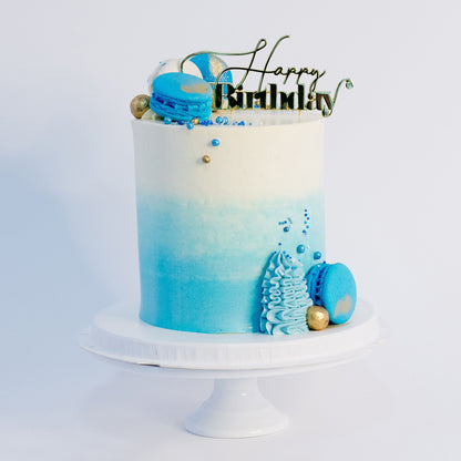 A beautifully decorated brithday cake cake with ombre blue frosting- buttercream, topped with white and blue macarons, gold maltesers, and a medley of blue, white, and gold sprinkles.Made be Cute Cakes and Co. Brisbane Cakes shop near you 