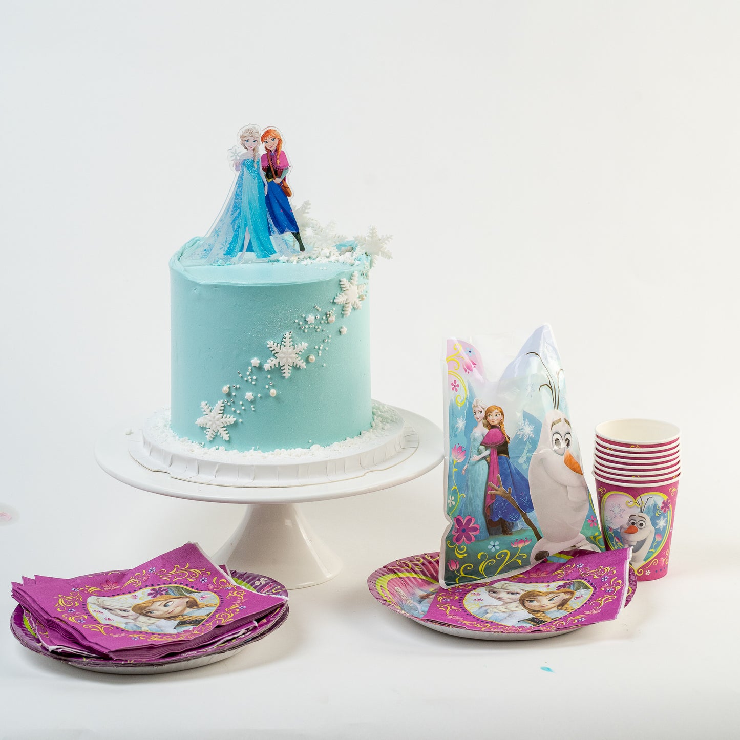 Frozen-themed birthday cake with decorations, cups, and plates on a white background