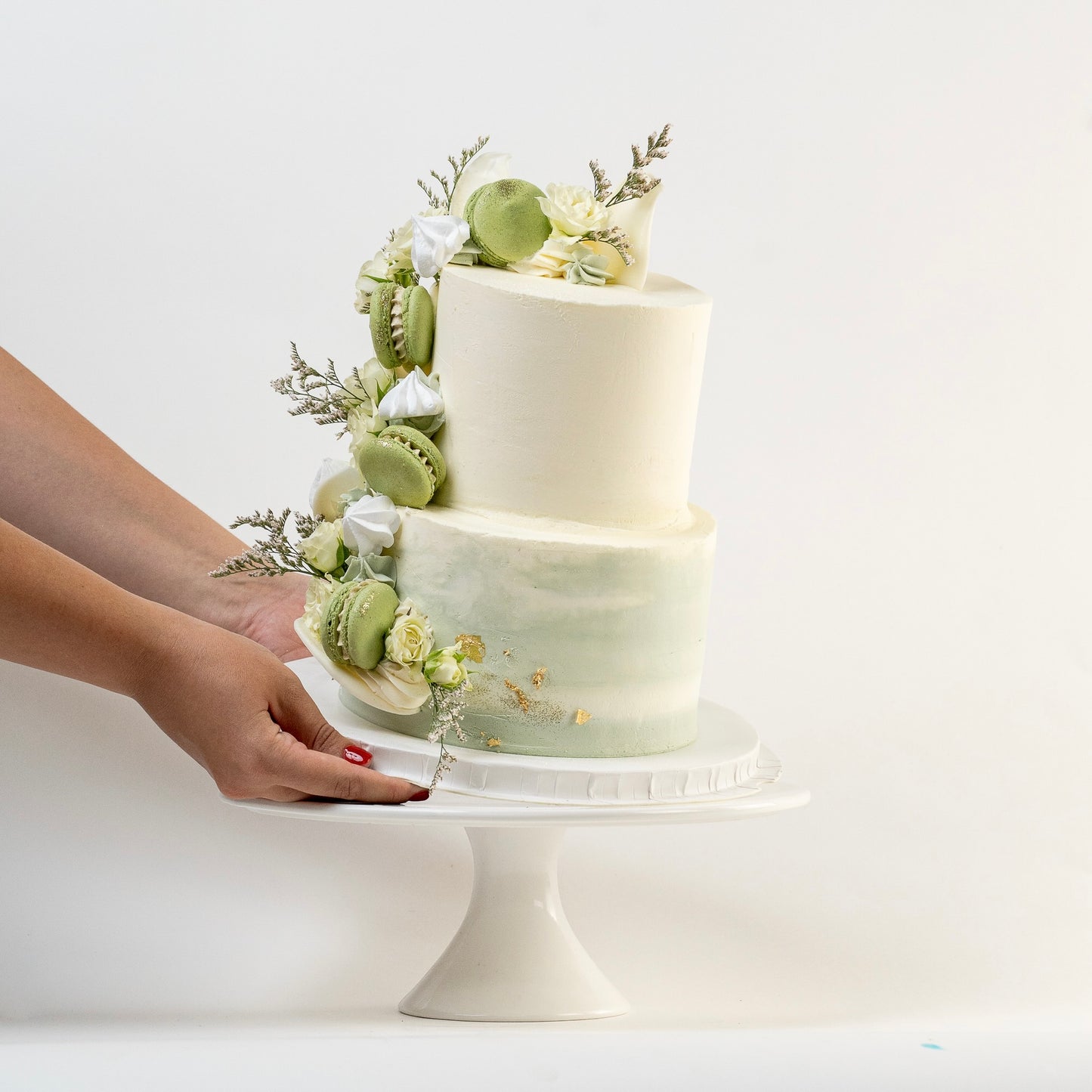 Two-tiered cake with green and white decorations on a white stand, held by a hand.
