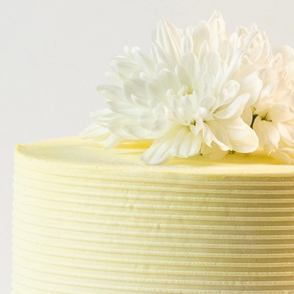 Close up image of a Light sherbet yellow colour cake. Decorated with ribbed buttercream feature on the sides and white fresh flowers on the top. Made with love by Brisbane’s favourite, 5 star cake shop, Cute Cakes & Co 