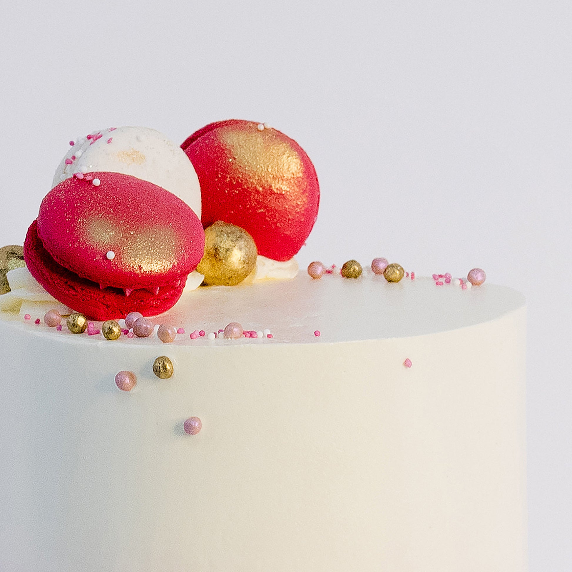A beautifully decorated birthday cake with  pink and white macarons, gold maltesers, and pink, white, and gold sprinkles. Made by Cute Cakes & Co . Brisbane 5 star rated cake shop that delivers the same day. 