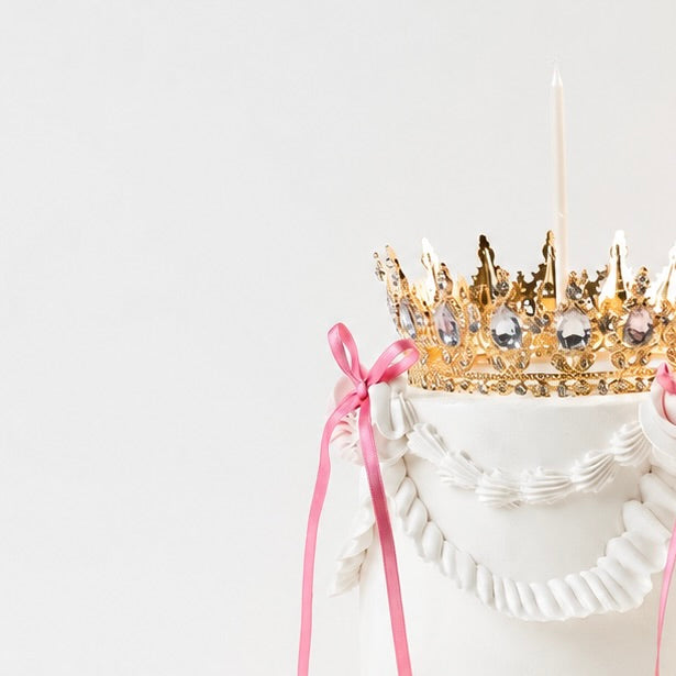 A lit candle on a white Vintage style birthday cake. Pink ribbon bow with a gold crown on the top
