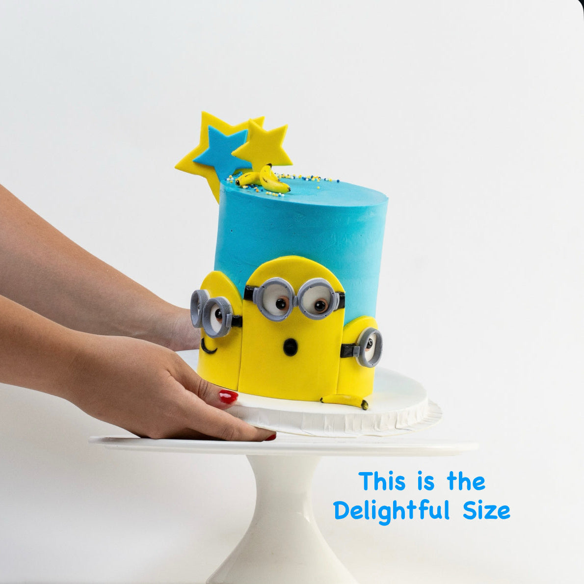 Minion birthday cake. with hand holding to so the size of the cake. Decorated with blue buttercream, three Despicable Me minions  characters on the front with icing bananas and stars as decoration.  Looks fabulous, Made with love by the five star cake shop ,Cute Cakes and Co, Brisbane. 