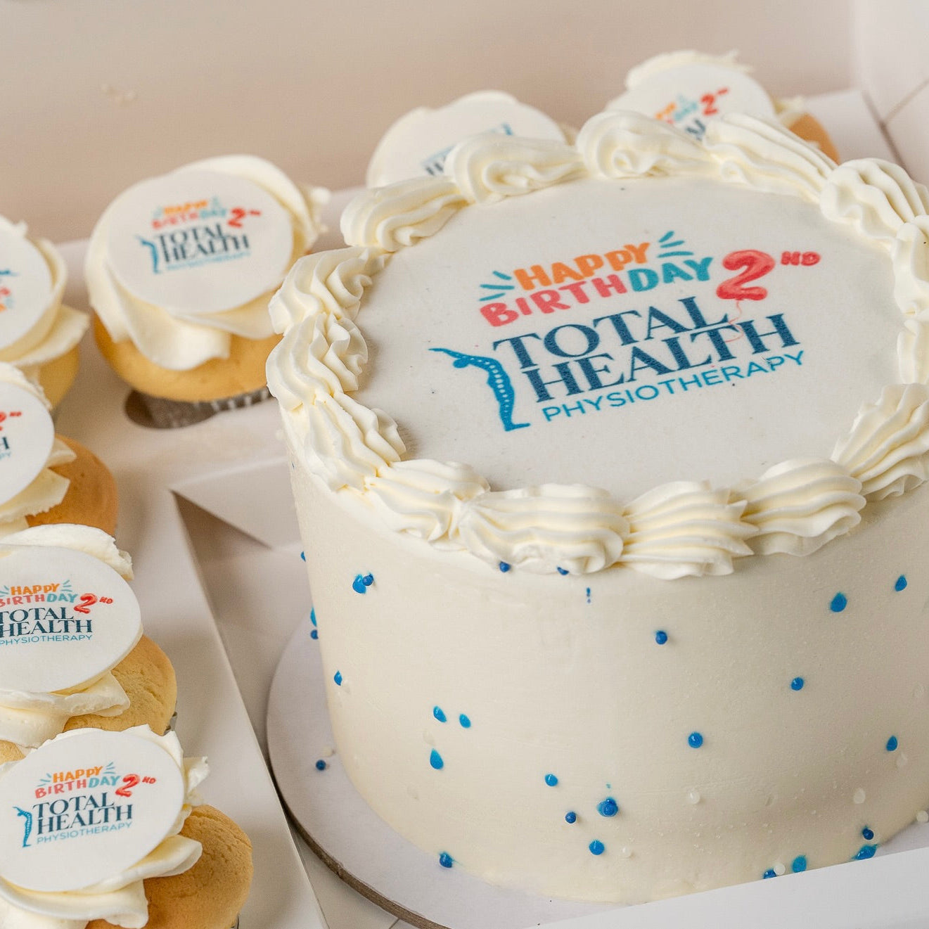 A edible image corporate celebration cake  cake with piped cream borders and blue sprinkles, topped 'Happy Birthday 2nd Total Health Physiotherapy.' Several matching cupcakes with identical toppers are arranged beside it in a box.