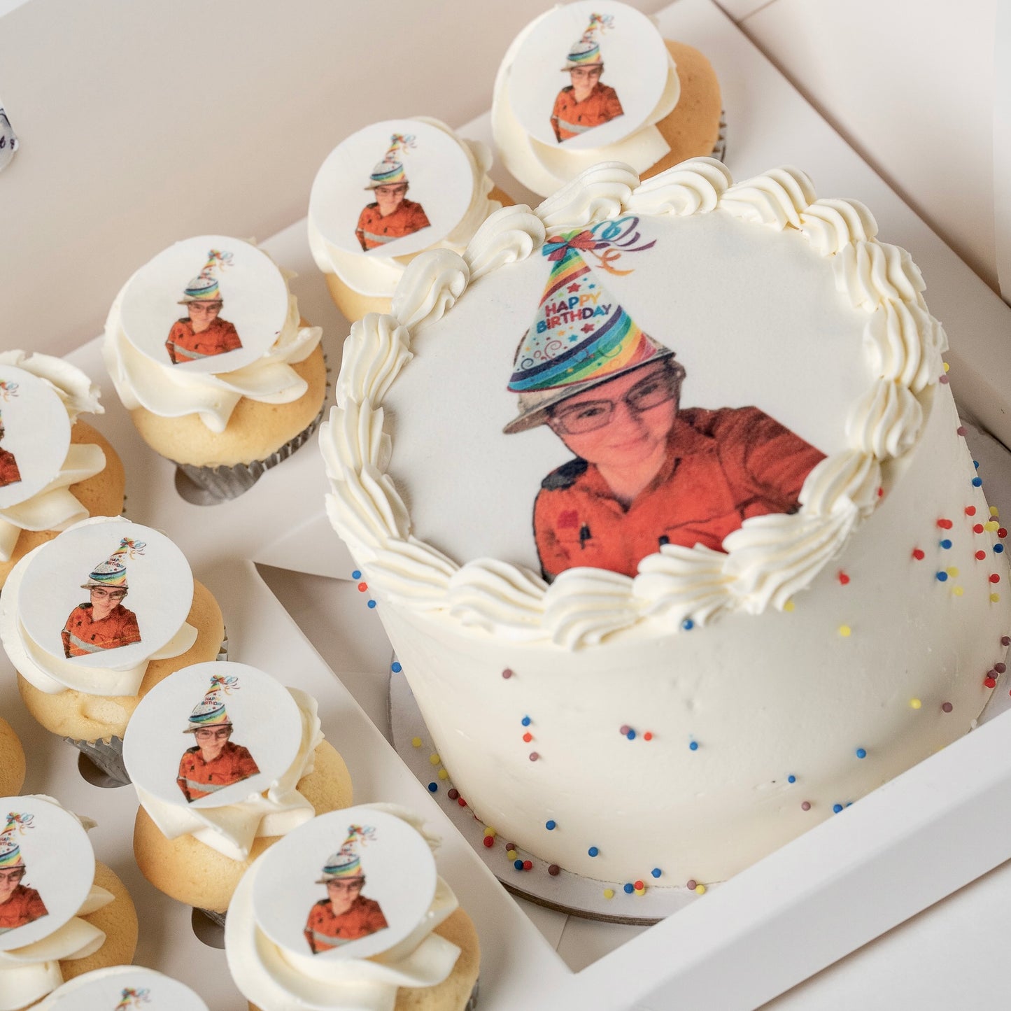 A white buttercream birthday cake and matching cupcakes in a box, each topped with a custom printed photo of a staff member and scattered colourful sprinkles.