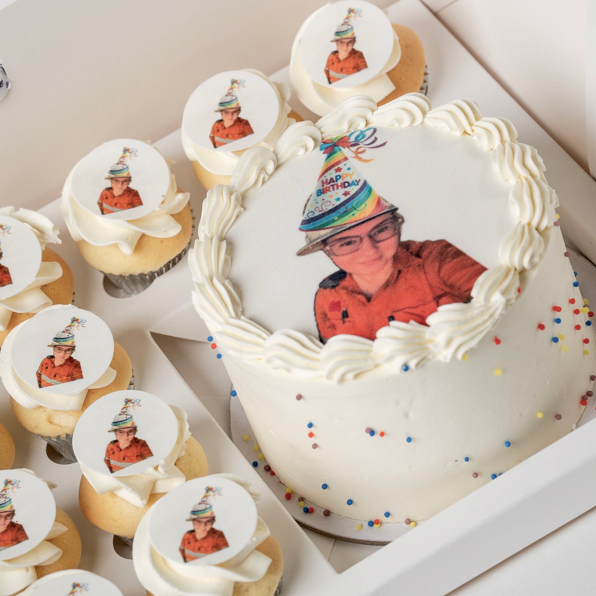 A white buttercream birthday cake and matching cupcakes in a box, each topped with a custom printed photo of a staff member and scattered colourful sprinkles.