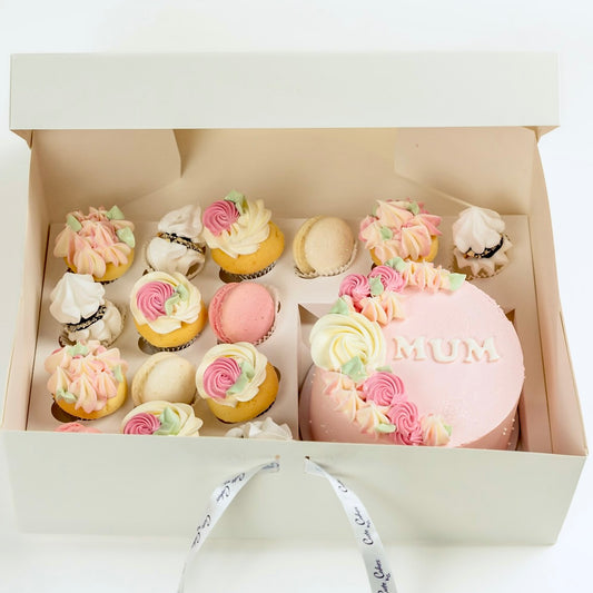 Decorative cake with 'MUM' on top, surrounded by smaller cupcakes in a box.