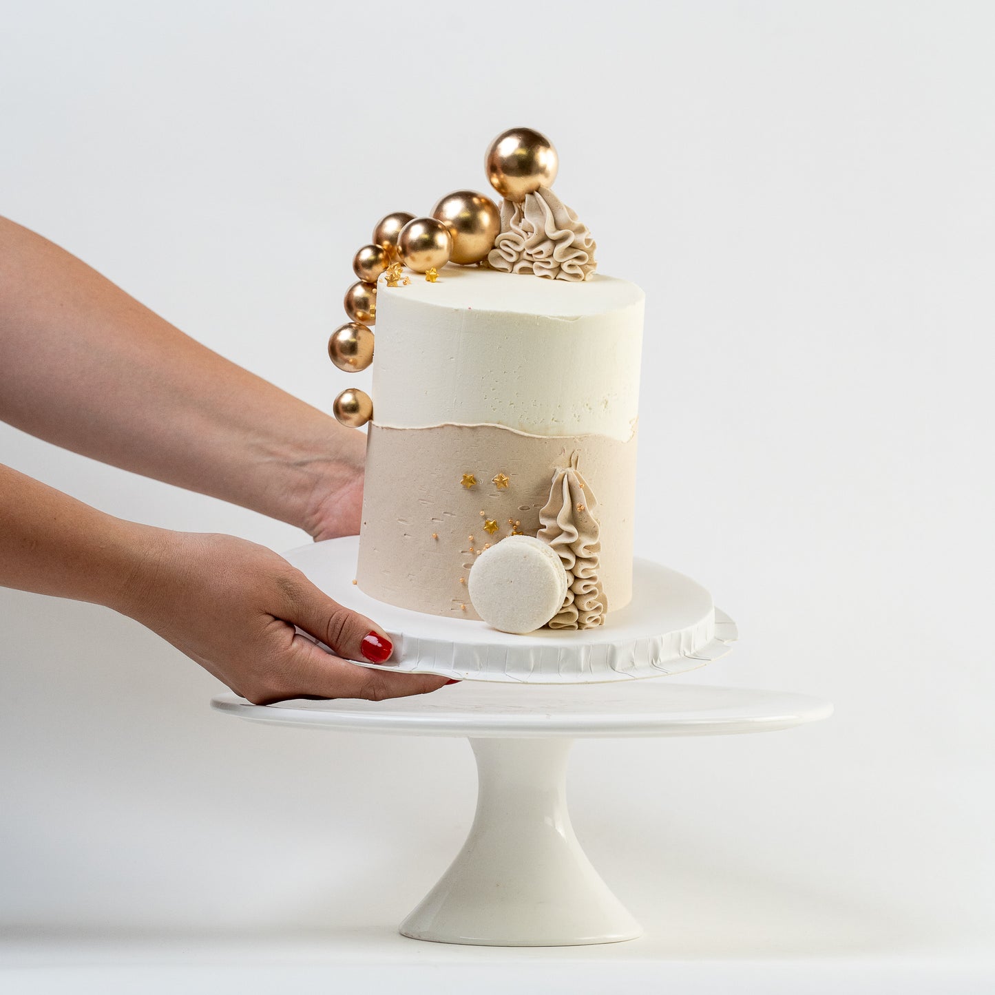 Hands placing down a white, caramel cake with gold balls and stars. Wiggle of buttercream at base and white Macaron. For perfect for engagements baby shower birthdays Looks fabulous, Made with love by Cute Cakes and Co. the 5 star review cake shop, in Brisbane.