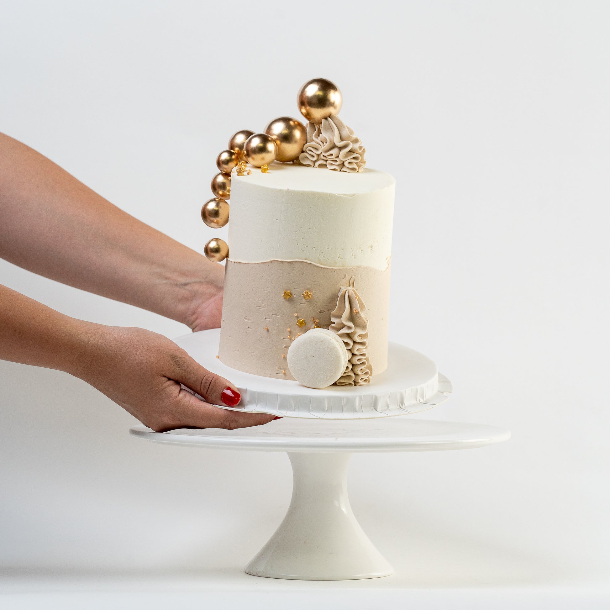 Hands placing down a white, caramel cake with gold balls and stars. Wiggle of buttercream at base and white Macaron. For perfect for engagements baby shower birthdays Looks fabulous, Made with love by Cute Cakes and Co. the 5 star review cake shop, in Brisbane.