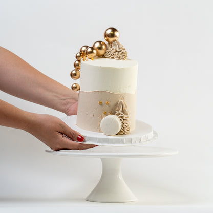 Hands placing down a white, caramel cake with gold balls and stars. Wiggle of buttercream at base and white Macaron. For perfect for engagements baby shower birthdays Looks fabulous, Made with love by Cute Cakes and Co. the 5 star review cake shop, in Brisbane.