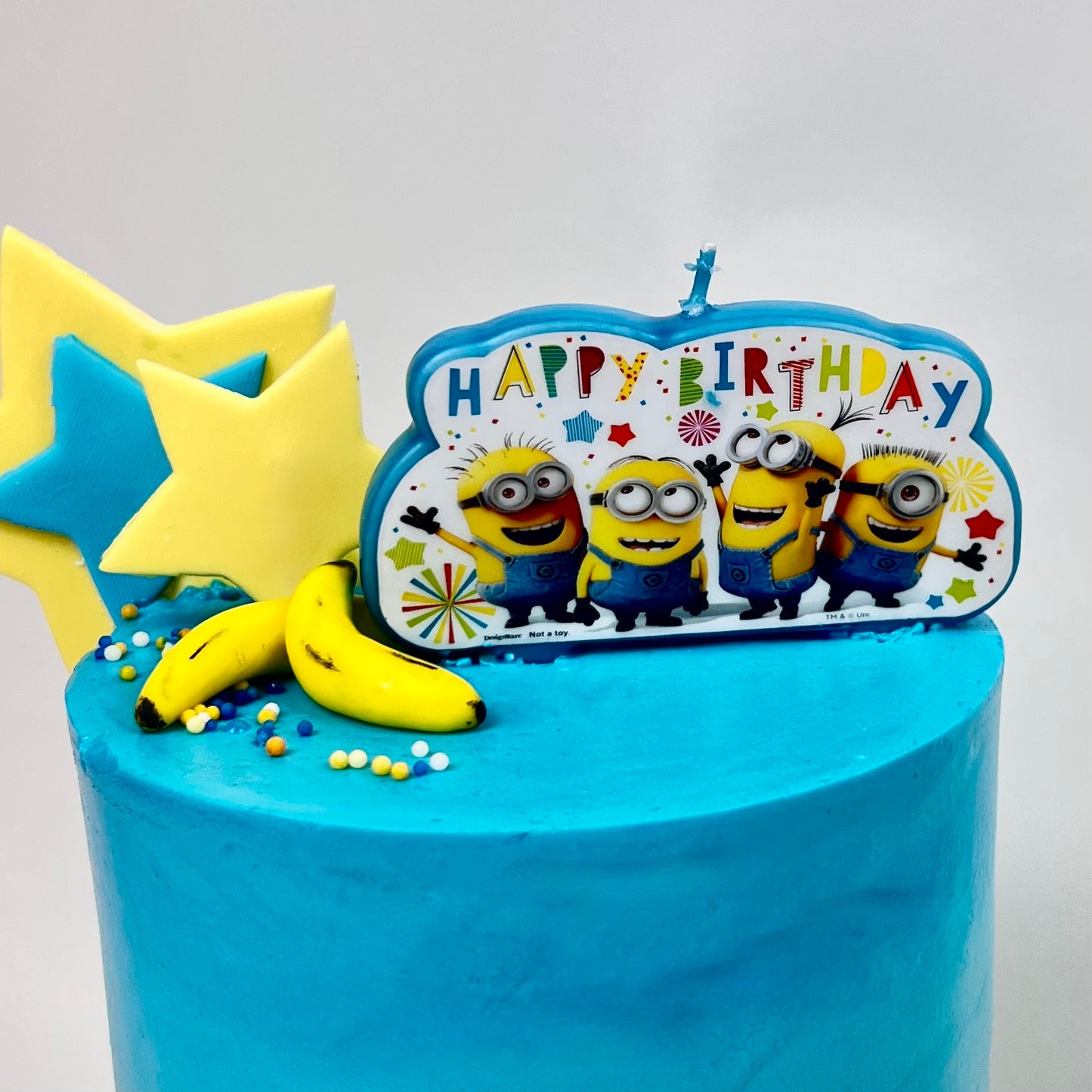 Blue birthday cake with Minion-themed 'Happy Birthday' candle on a white background
