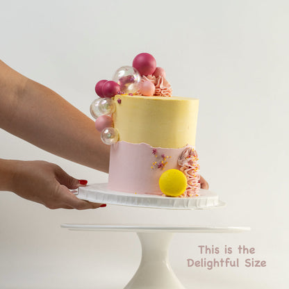 Hands placing down a pretty pink and yellow cake with a side decoration of shades of pink and clear balls. Wiggle and yellow macaron at the base. glitter perfect for a  girl's birthday Looks fabulous.  Made with love by Cute Cakes & Co, Brisbane Favourite cake shop. 
5 star review cake shop in Brisbane. 
