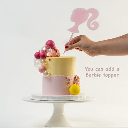 Hands placing down a Barbie head cake topper on the top of  Pretty pink and yellow cake with a side decoration of shades of pink and clear balls. Wiggle and yellow macaron at the base. glitter perfect for a  girl's birthday Looks fabulous.  Made with love by Cute Cakes & Co, Brisbane Favourite cake shop. 
5 star review cake shop in Brisbane. 