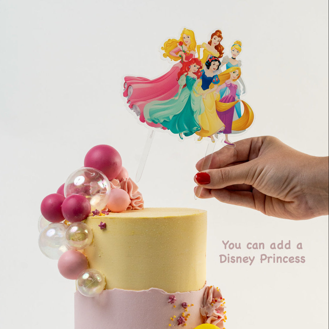 Hands placing down a Disney Princess cake topper on the top of  Pretty pink and yellow cake with a side decoration of shades of pink and clear balls. Wiggle and yellow macaron at the base. glitter perfect for a  girl's birthday Looks fabulous.  Made with love by Cute Cakes & Co, Brisbane Favourite cake shop. 
5 star review cake shop in Brisbane. 