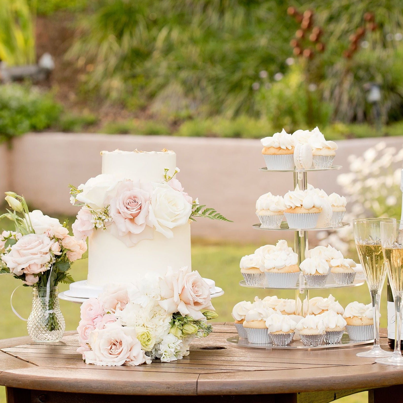 Outside wedding scene with a beautiful Wedding cake with flowers, cupcake stand and champagne on a wooden table outdoors.