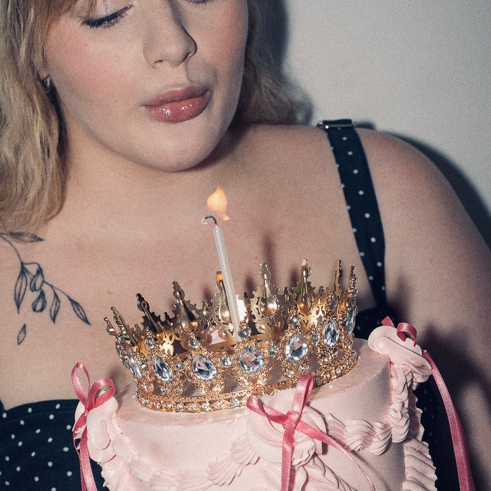A woman blowing out a candle on a pink Vintage style birthday cake. Pink ribbon bows with a gold crown on the top and candle. Made with love by Brisbane’s favourite, 5 star cake shop, Cute Cakes & Co 